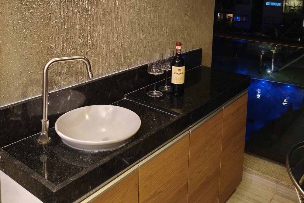 Varanda gourmet à noite, com bancada de granito, cuba e a piscina iluminada ao fundo