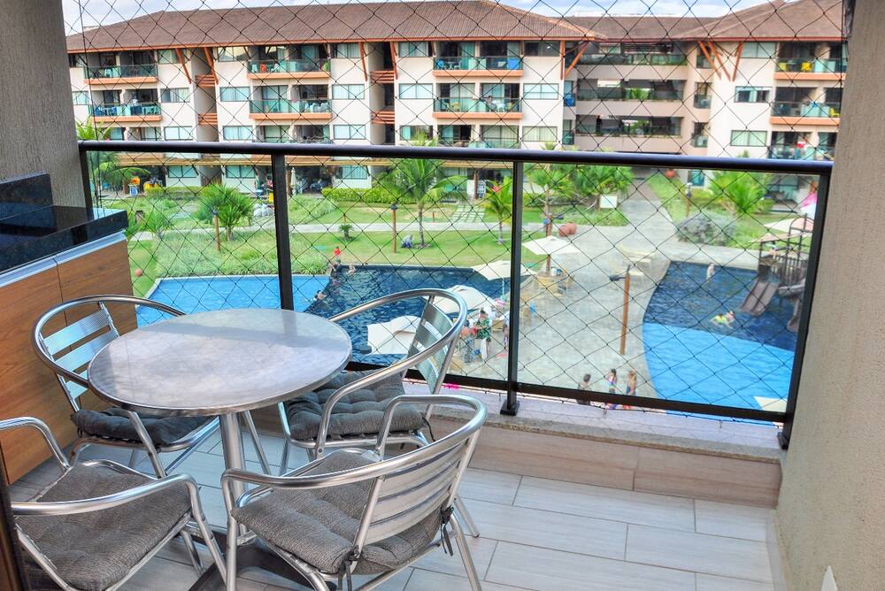 Varanda do apartamento com mesa, tela de proteção e vista para as piscinas